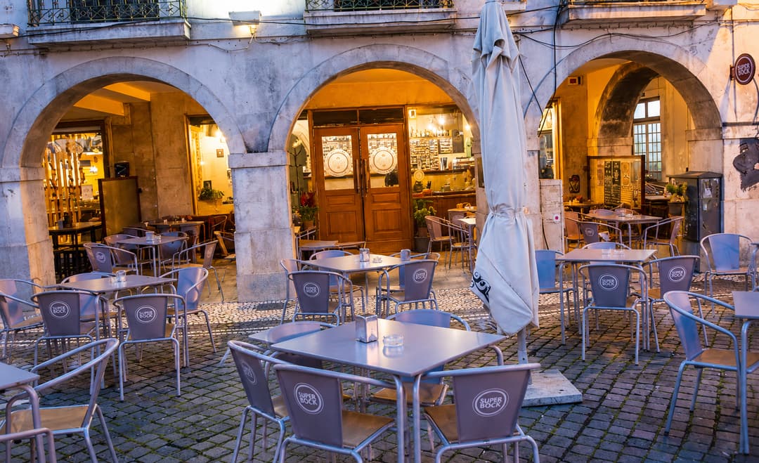 Esplanada do Praça Caffè sob as arcadas de pedra, ao anoitecer, com mesas na calçada e o interior iluminado por trás das portas de madeira.