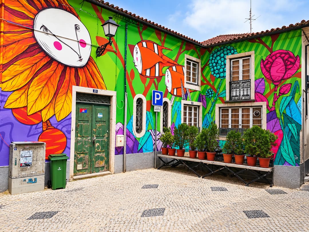 Fachada coberta por um mural de arte urbana muito colorido, com uma grande flor laranja de rosto pintado, peixes e flores, junto a uma rua de calçada.
