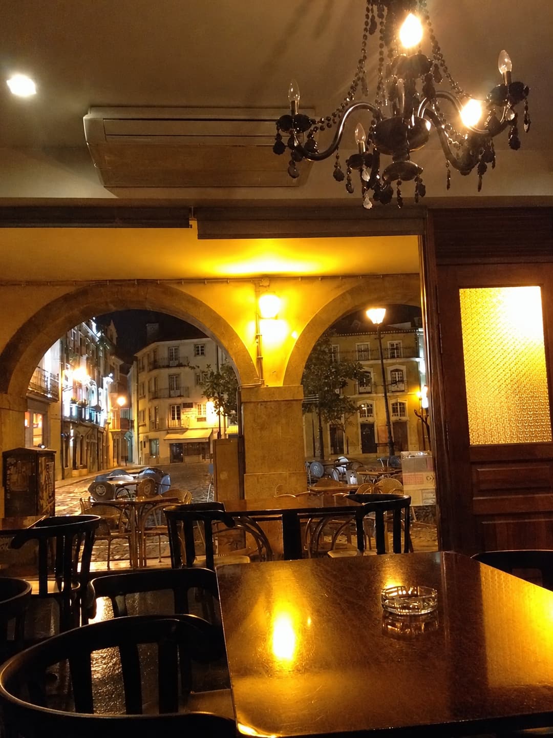 Interior do Praça Caffè à noite: lustre de cristal negro, mesa de madeira escura em primeiro plano e, através das arcadas, a praça iluminada.