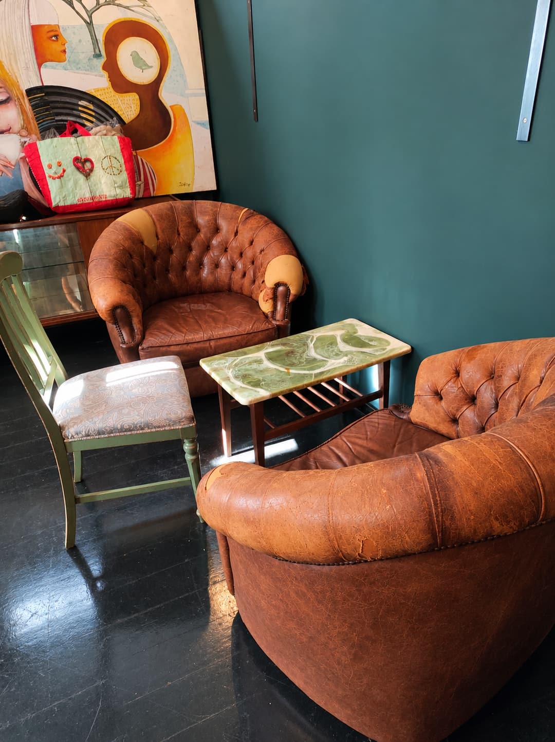 Recanto com dois cadeirões capitoné em pele castanha, uma mesa baixa de ónix verde e uma cadeira de madeira pintada de verde, junto a uma parede verde-petróleo.