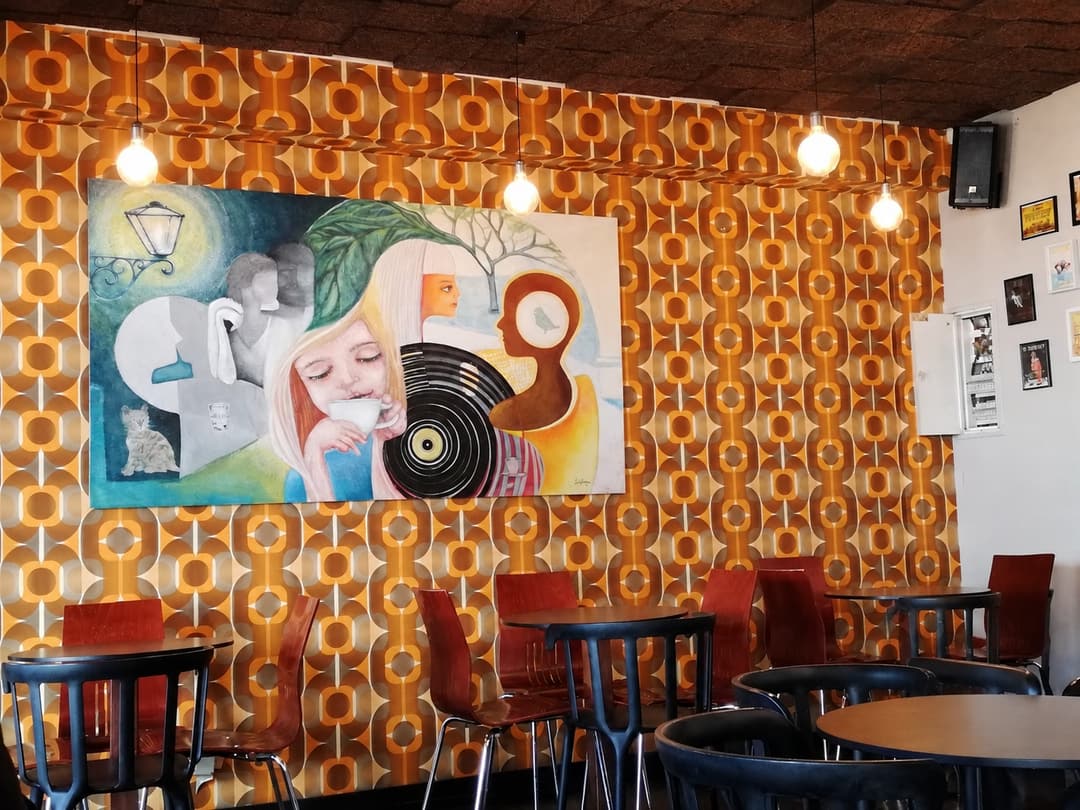 Sala com papel de parede retro em tons de laranja e castanho, lâmpadas suspensas e um grande quadro com uma mulher a beber café ao lado de um disco de vinil.