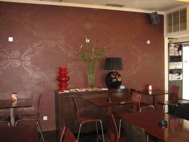 Sala com paredes grená ornamentadas com motivos florais dourados, mesas e aparador em madeira escura, uma jarra vermelha e um candeeiro preto.